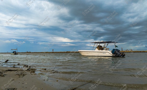 A white fishing boat on the water near the shore, equipped with a large outboard motor. The background features a cloudy sky with a distant horizon line. Small rocks and sand are visible in the foreground on the shore.