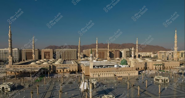 Aerial view of the Prophet\'s Mosque in Medina, showcasing the expansive courtyards, minarets, surrounding buildings, and mountains in the background.