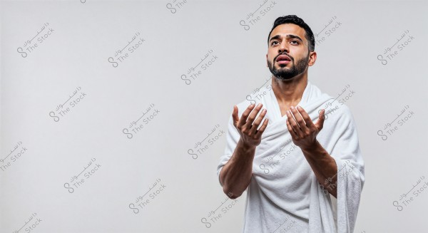 Image of a man wearing a white Ihram, standing with his hands raised towards the sky in a prayer position. The background is plain and white, emphasizing the individual in the photo. The man appears to be in a state of contemplation and devotion.