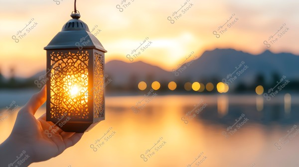 A hand holding a traditional lantern with geometric patterns, illuminated with a warm glow against a sunset backdrop. The background features a lake and mountains, with hues of orange and gold.