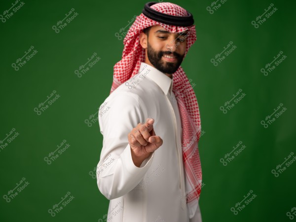 A man wearing a Saudi thobe and ghutra stands in front of a green background, pointing with his hand.