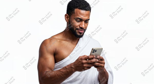 A portrait of a man holding a phone and smiling while looking at it. The man is wearing a white towel wrapped around his bare shoulder, and he appears to be standing in a well-lit environment with a plain white background.