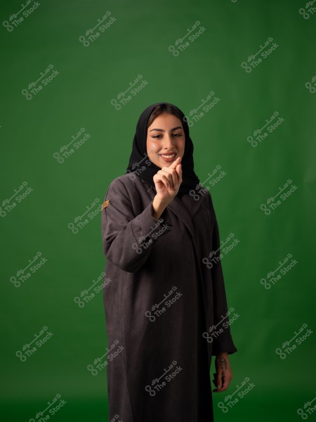 صورة لامرأة ترتدي عباية سوداء وحجاب أسود واقفة أمام خلفية خضراء. تظهر المرأة مبتسمة وهي تشير بيدها اليمنى نحو الأسفل, مرتدية عباية داكنة اللون.