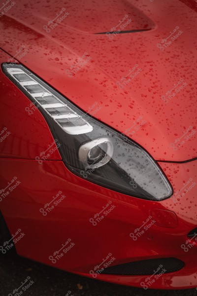 Close-up of a red car\'s front headlight covered with raindrops, giving a glossy appearance to the paint.