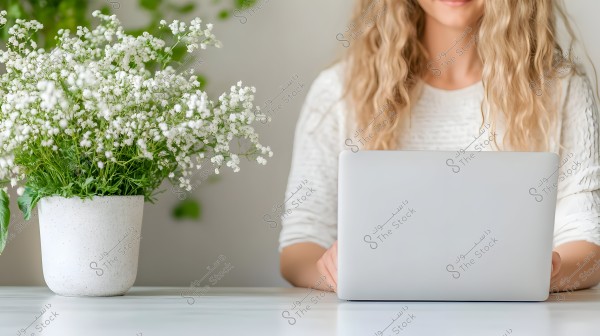 An image of a woman sitting and working on a laptop. She is wearing a white top. To her left, there is a white vase containing small white flowers. The background is neutral with soft green details.