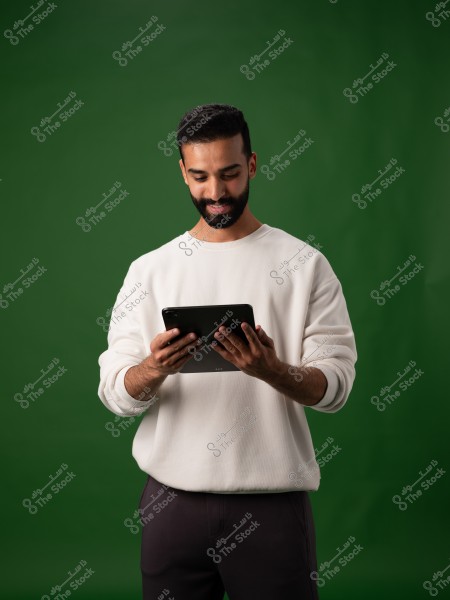 A portrait of a bearded man wearing a white sweater and dark pants, standing in front of a green background. He is holding a tablet in his hands and looking at it with a slight smile.
