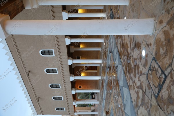 An image of a traditional building featuring mud facades and white columns. The building has arched windows and a decorative roof. The floor is paved with various colored stones, and some lights are illuminated, adding a heritage atmosphere to the setting.