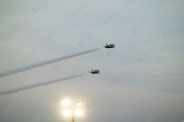 The image shows two fighter jets flying in the sky, leaving trails of smoke behind. The jets appear in green and white colors, flying in a streamlined formation. At the bottom of the image, there are bright lights, indicating that the air show is taking place in the early evening or at sunset.