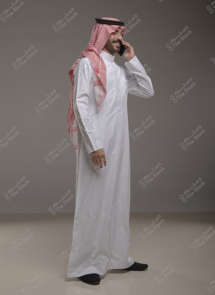 A portrait of a man standing and wearing a white thobe and a red and white checked ghutra with a black agal, while speaking on a mobile phone in a simple studio setting with a grey background. The attire is traditional and suggests it is from the Gulf countries, such as Saudi Arabia.