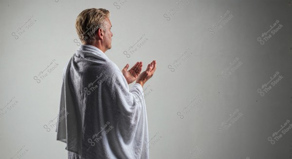 A side view image of a man wearing a white Ihram, possibly engaged in Hajj or Umrah rituals. The man is shown in a prayer position with hands raised. The background is neutral gray, highlighting the clothing details and soft lighting on the individual.