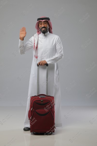 The image shows a man standing and wearing a traditional white Saudi thobe with a red and white shemagh and a black agal. He holds a large red wheeled suitcase by its handle while raising his other hand in a friendly wave with a smile. The man is in a studio setting with a gray background.