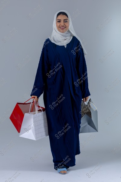 An image of a woman wearing a blue abaya and a white hijab, holding multiple colorful shopping bags. She appears happy and stands against a simple gray background. The clothing suggests a Middle Eastern style.