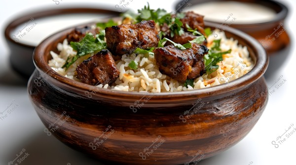 An image showing a wooden bowl filled with white rice garnished with pieces of grilled meat and green cilantro leaves. In the background, there are other wooden bowls with blurred side dishes.