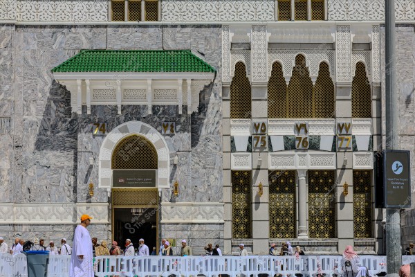 Image of an entrance to the Masjid al-Haram in Mecca, showcasing an ornate architectural design with marble and Islamic motifs. Above the door are plaques with gate numbers: 74, 75, 76, and 77. Several people are seen in the foreground, most wearing traditional attire such as white robes and headscarves. A white plastic barrier is in front, and there is a sign indicating escalators leading to upper floors.