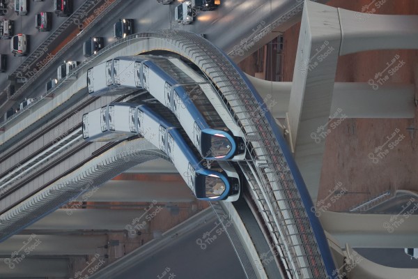 An aerial view of two trains moving in opposite directions on an elevated track next to a busy highway with cars. Each train is blue and white and travels on a curved track. The highway is crowded with cars moving in parallel lines beneath the trains.