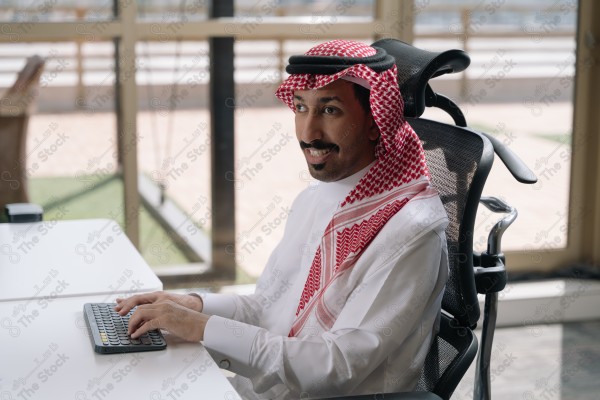 رجل سعودي "من ذوي الإعاقة" مبتسم يرتدي الثوب السعودي التقليدي ويجلس على كرسي مكتب ويستخدم جهاز للكتابة.