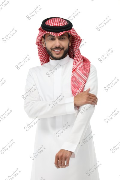 Portrait of a man wearing traditional Saudi attire, featuring a white thobe and a red checkered ghutra with a black agal. The man is smiling and has his hand on his arm. The background is entirely white.