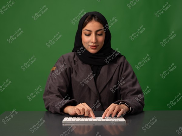 امرأة ترتدي حجابًا أسود وجلابة رمادية اللون تجلس أمام خلفية خضراء, وتستخدم لوحة مفاتيح على طاولة.
                            
-
