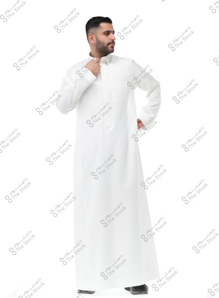 Image of a man wearing a white thobe, standing against a white background. He is dressed in traditional Gulf attire, with short hair and a beard. The white thobe is simple and elegant, and the man is seen adjusting the collar of the thobe with his hand.