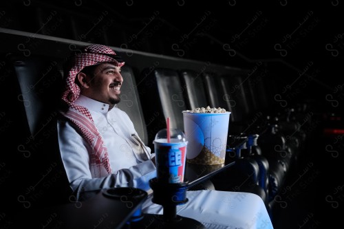 شاب سعودي يرتدي الزي السعودي التقليدي يجلس في صالة السينما ويظهر عليه السرور أثناء مشاهدة الفيلم، رجل سعودي يرتدي الزي السعودي التقليدي يجلس في صالة السينما ويظهر أمامه الفشار وأحد المشروبات على الطاولة