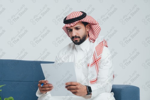 رجل سعودي يرتدي الزي السعودي التقليدي يجلس على أريكة ويراجع ورقة عمل