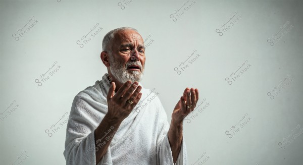 Image of an elderly man wearing white Ihram clothing, raising his hands in prayer, with a serene expression on his face. The background is plain and smooth.