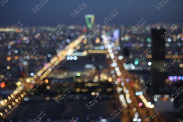 A blurred image of a large city at night, with twinkling city lights in the background. The roads are brightly lit with various colorful lights. In the background, a distinct skyscraper with a unique structure can be seen, likely the Kingdom Centre in Riyadh, Saudi Arabia.