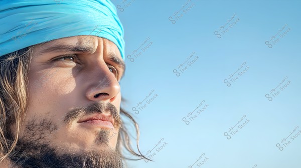 صورة لشخص ينظر إلى البعيد تحت سماء زرقاء صافية. يبدو مرتديًا عمامة أو وشاحًا أزرق على رأسه. يظهر جزء من وجهه ولديه لحية قصيرة وشارب, وتبدو تعابيره جادة ومركزة.