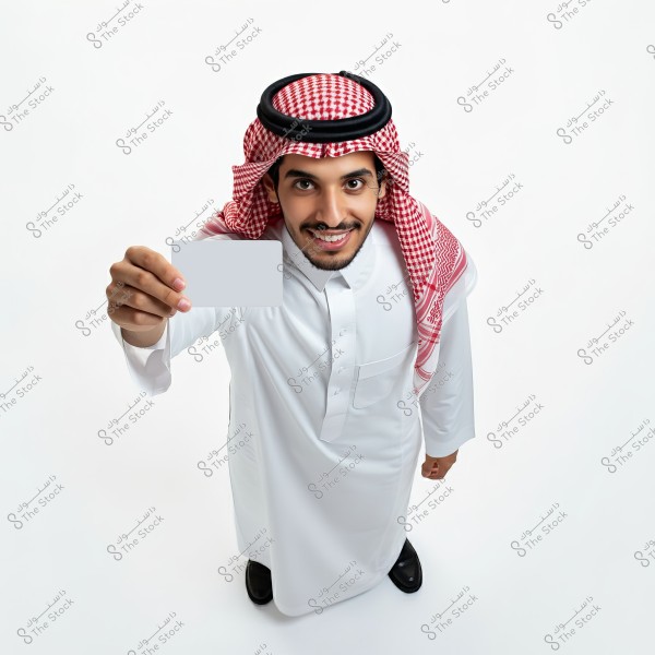 A man wearing a white thobe and a red checkered headscarf, smiling and holding a blank white card in his hand. The photo is taken from an overhead angle with a white background.