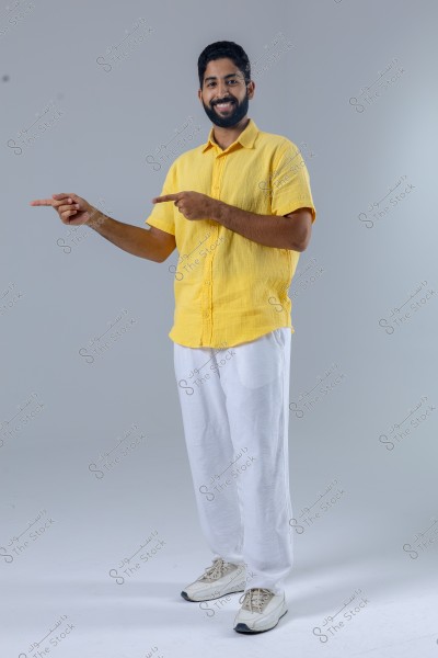 A portrait of a man standing, smiling, and pointing his fingers to the left. He is wearing a yellow shirt, white trousers, and white sneakers. The background is plain white, highlighting the person in the image.