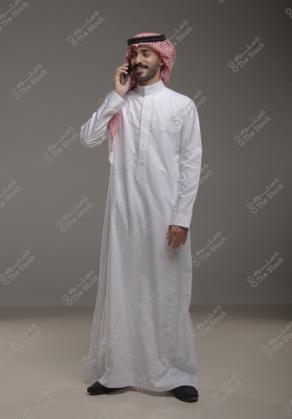 A portrait of a man wearing a white thobe, red checkered ghutra, and a black agal, talking on his mobile phone. He appears happy and stands against a grey background. The clothing suggests traditional Saudi attire.