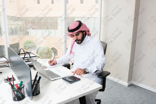 رجل سعودي يرتدي الزي السعودي التقليدي يعمل في مكتب بواجهة زجاجية ويستخدم قلم رصاص لتدوين الملاحظات