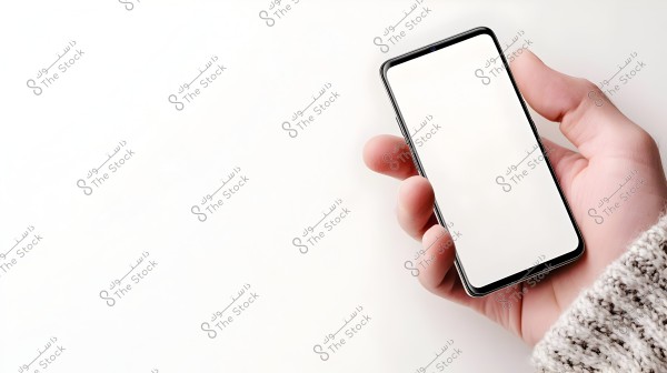 Image shows a person’s hand holding a smartphone with a blank white screen. The hand is wearing a knitted sweater with sleeves in gray tones. The background is white, highlighting the phone and hand.