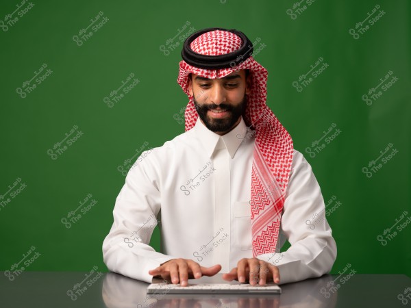 رجل يرتدي الزي السعودي التقليدي, جالس أمام حاسوب ويمسك لوحة مفاتيح على طاولة. يرتدي ثوبًا أبيض وغترة حمراء وبيضاء بينما يظهر تعبير وجهه هادئًا. الخلفية خضراء اللون.

-