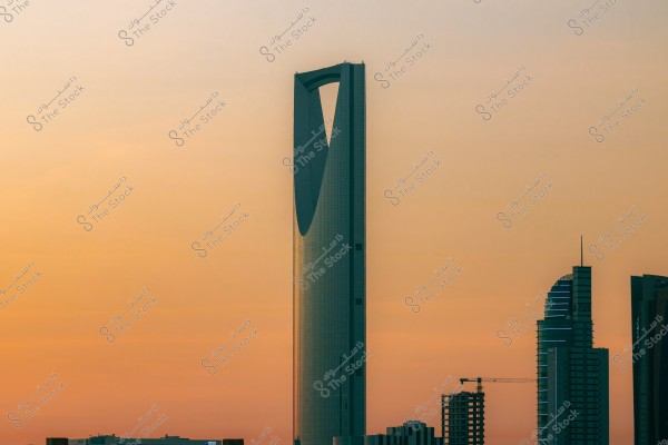 The image shows the Kingdom Tower in Riyadh, Saudi Arabia at sunset, with a sky filled with warm tones. The tower is surrounded by various tall buildings in the city skyline.