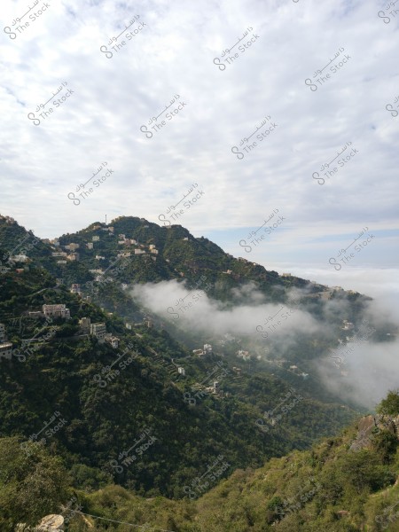 صورة لمنظر طبيعي يظهر سلسلة من الجبال المغطاة بالخضرة الكثيفة. تتخلل الضباب أجزاء من الجبال وتغطيها الغيوم البيضاء. تنتشر بعض المباني على سفوح الجبال, مما يعطي انطباعًا لمجتمع سكني محلي في منطقة جبلية. السماء مغطاة بالغيوم بشكل جزئي.