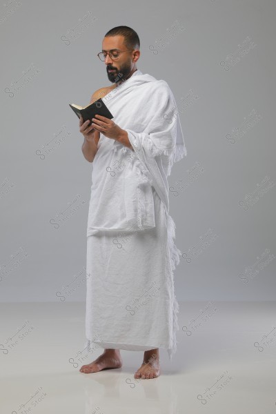 Image of a man wearing a white Ihram, holding a small open book in his hands. He is standing barefoot on a white floor, with a grey background. He appears to be focused on reading.
