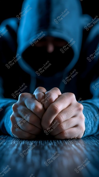 An image depicting a person wearing a hooded sweatshirt, with their face concealed in shadow. The hands are clasped together on a wooden table, appearing contemplative. The lighting is dim, creating a mysterious and tranquil atmosphere.