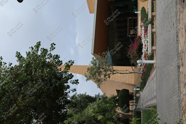 Image of a modern architectural building facade, featuring a combination of glass and wood design, surrounded by a neatly manicured garden with colorful flowers. In the foreground, there is a stone walkway lined with green trees and blooming plants.