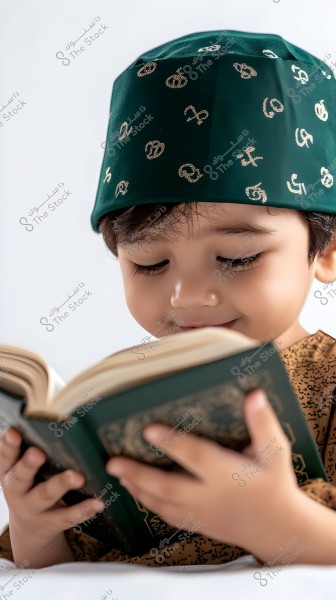 A portrait of a young child sitting and attentively reading a book. The child is wearing a decorative green cap and a traditional brown-patterned outfit, suggesting a possible Arabic cultural origin. The green cap is adorned with golden symbols.