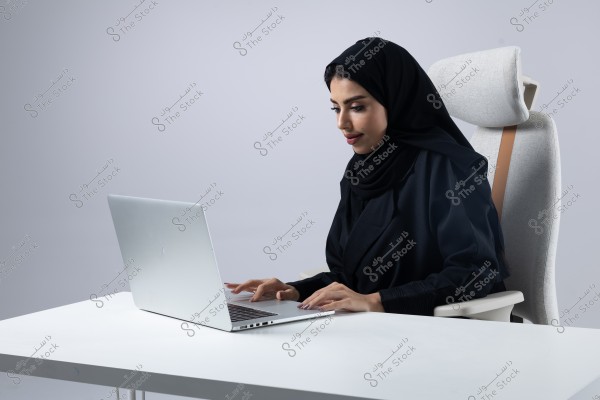 The image shows a woman sitting on an office chair and working on a silver laptop placed on a white table. She is wearing a black hijab and a black abaya, focusing intently on the laptop screen. The background is plain white and calm.