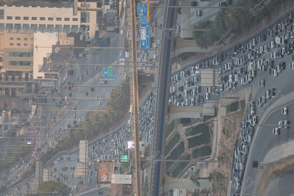 Aerial view of traffic congestion in a modern city, showcasing a large number of vehicles halted across several lanes on a main road, with a flyover connecting the different directions. Tall city buildings are visible in the background, along with palm trees lining the road. There are blue directional signs with Arabic writing, and the sky appears slightly overcast.