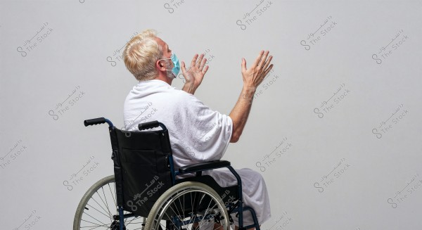 A man sitting in a wheelchair, wearing a white robe and a blue medical face mask, raising his hands upwards as if in prayer or supplication, with a plain white background.