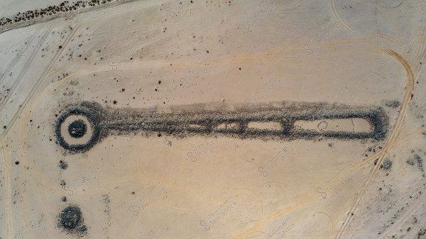 An aerial view of a circular and rectangular formation on the desert sands. The circular shape consists of a layer of dark stone surrounding a light-colored sandy circle, with a straight rectangular line extending from it. The formation is set amidst a wide sandy expanse with small scattered plants and tire tracks visible on the sand.