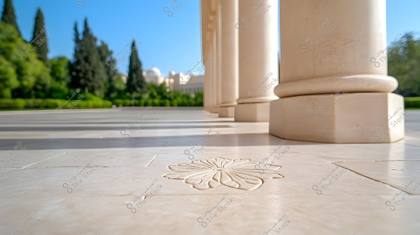 An image of large marble columns lined up in a row, with a flower design carved into the stone floor in the foreground. In the background, there is a view of green trees and a distant building wall. The sky is clear blue.