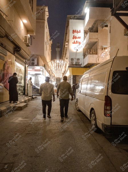 The image shows a narrow street at night in a Middle Eastern city, with decorative lighting hanging across the street. Two people are walking beside a white van parked on the right side. Buildings on both sides of the street have signs in Arabic, and several other people are visible in the background. A minaret is visible in the distance.