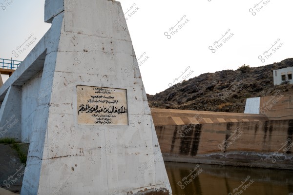 The image shows a large concrete structure with a commemorative plaque written in Arabic, indicating a project related to dams. In the background, natural hills and rocks are visible.