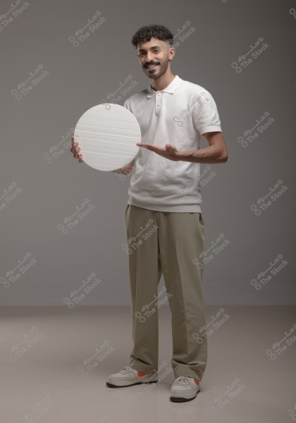 A man wearing a white polo shirt and beige pants stands in a gray studio. He is holding a white circle in his hand and pointing to it with a smile on his face. He is wearing white athletic shoes with an orange stripe.