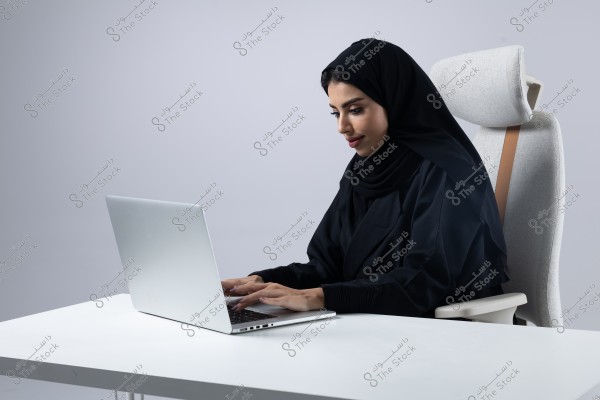 Image of a woman sitting on an office chair, wearing a black abaya and hijab, working on a laptop on a white desk. The background is simple with gray tones.