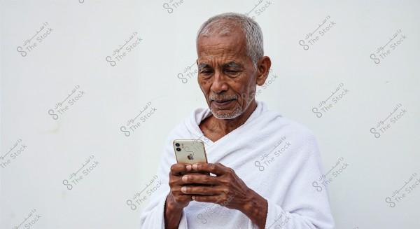 An image of an elderly man dressed in a white ihram garment, focused on a smartphone in his hands. The setting is neutral with a white wall background, suggesting he might be preparing for religious rituals like Umrah or Hajj.
